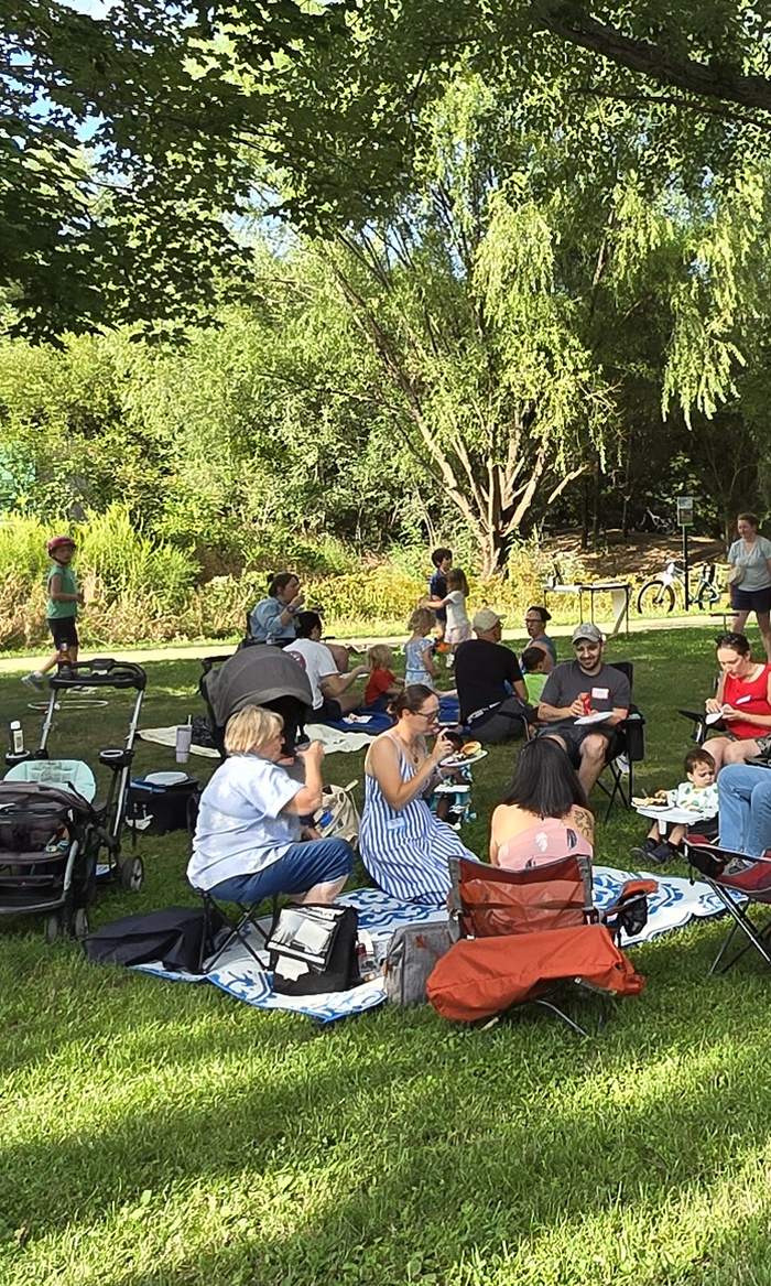 Neighbors sitting in camp chairs and on blankets under the shade trees at Evans Parkway Park during the Block Party
