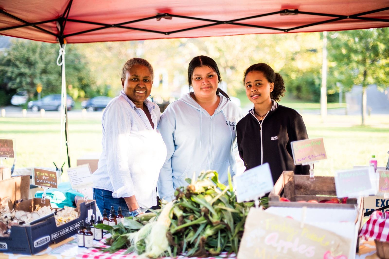 Three neighbors smiling behind a produce stand at the GNA booth at a local farmers market