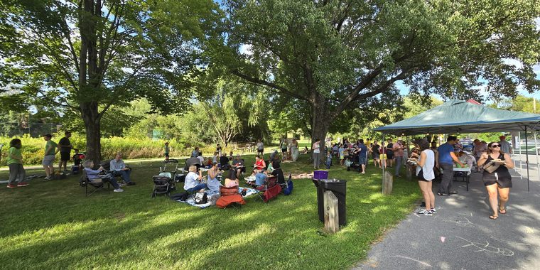 Neighbors sitting in camp chairs and on blankets under big shade trees, with a line of people at the food tent along the street