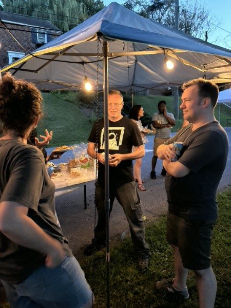 Three neighbors talking under a canopy at dusk with string lights overhead