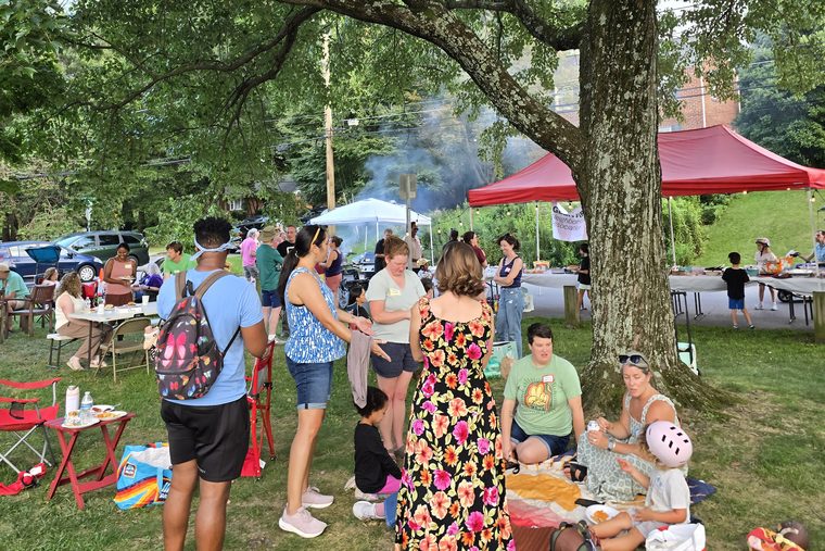 Neighbors standing and talking in groups while others sit on blankets, the red canopy and grill smoke behind them