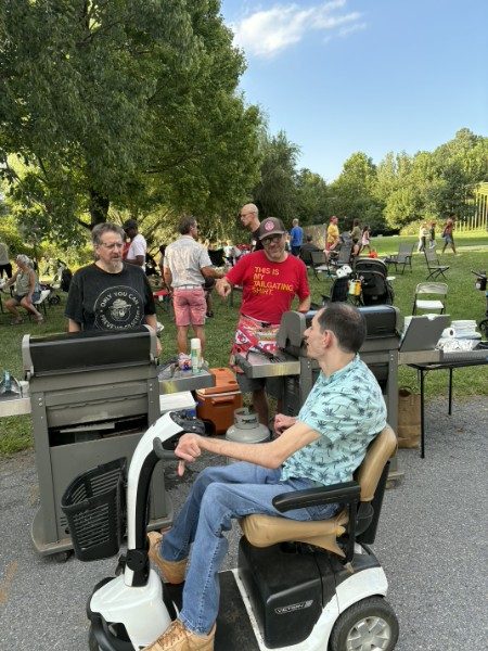 Two large grills on the pavement with a neighbor tending them, another neighbor on a mobility scooter alongside, and a crowd on the grass behind