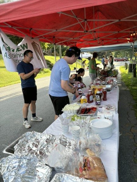 A long white table covered end to end with dishes, condiments and stacks of plates, with neighbors serving themselves